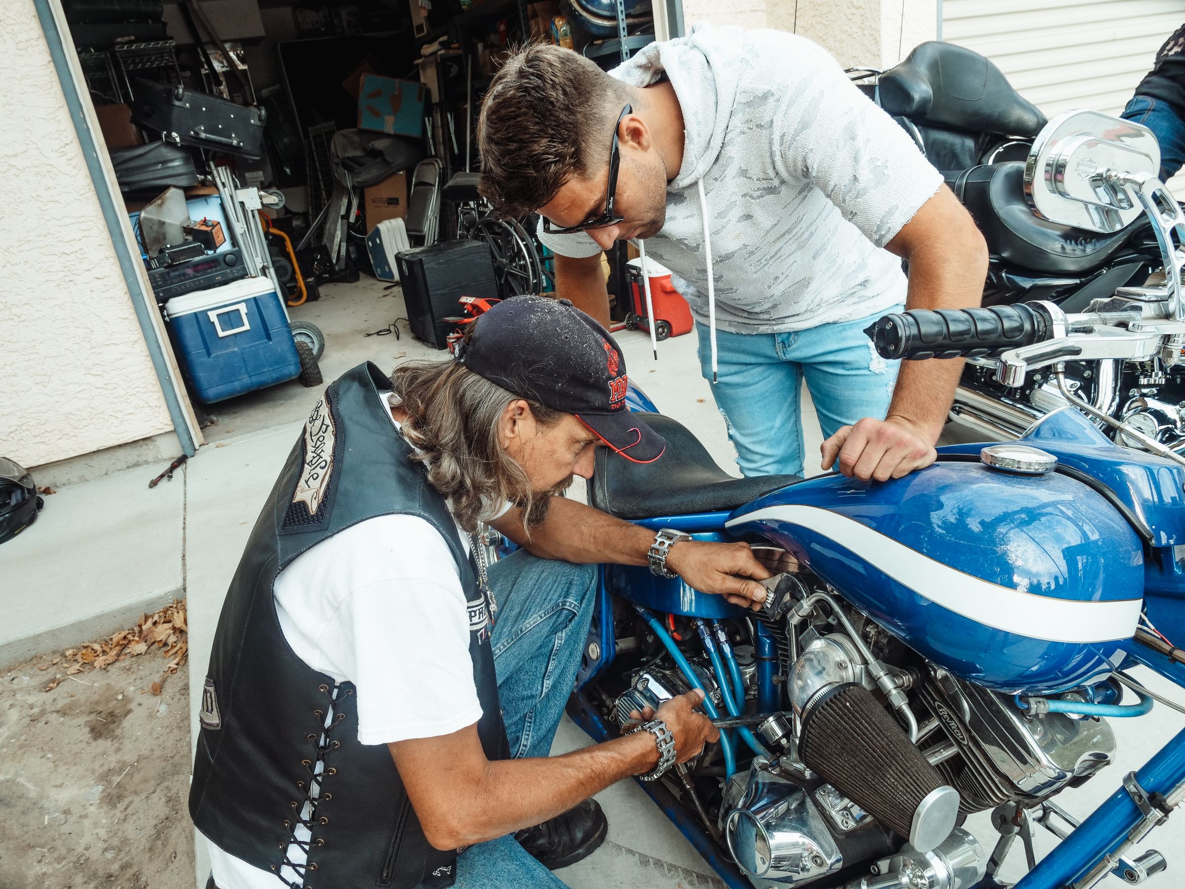 Two men repairing a motorcycle together in a cluttered garage, one teaching the other