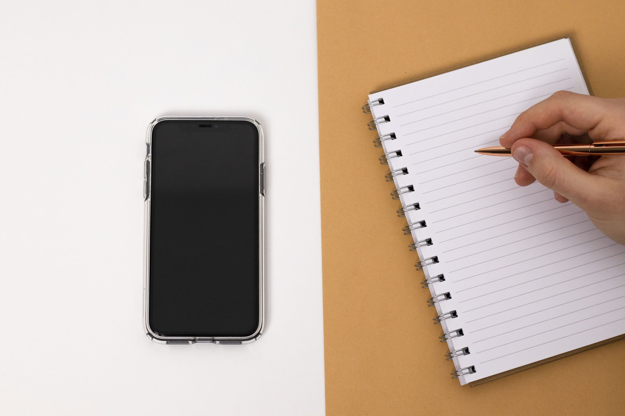 A phone and a notebook on a desk with a hand holding a pen, the quiet act of writing a review that outlives the evening it describes