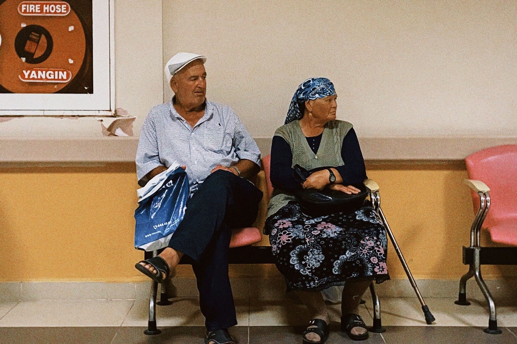 A couple sitting quietly in a hospital corridor waiting area, the people who absorb the lateness of others