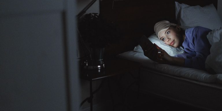 A young woman lying in bed at night, her face lit only by the glow of a phone screen