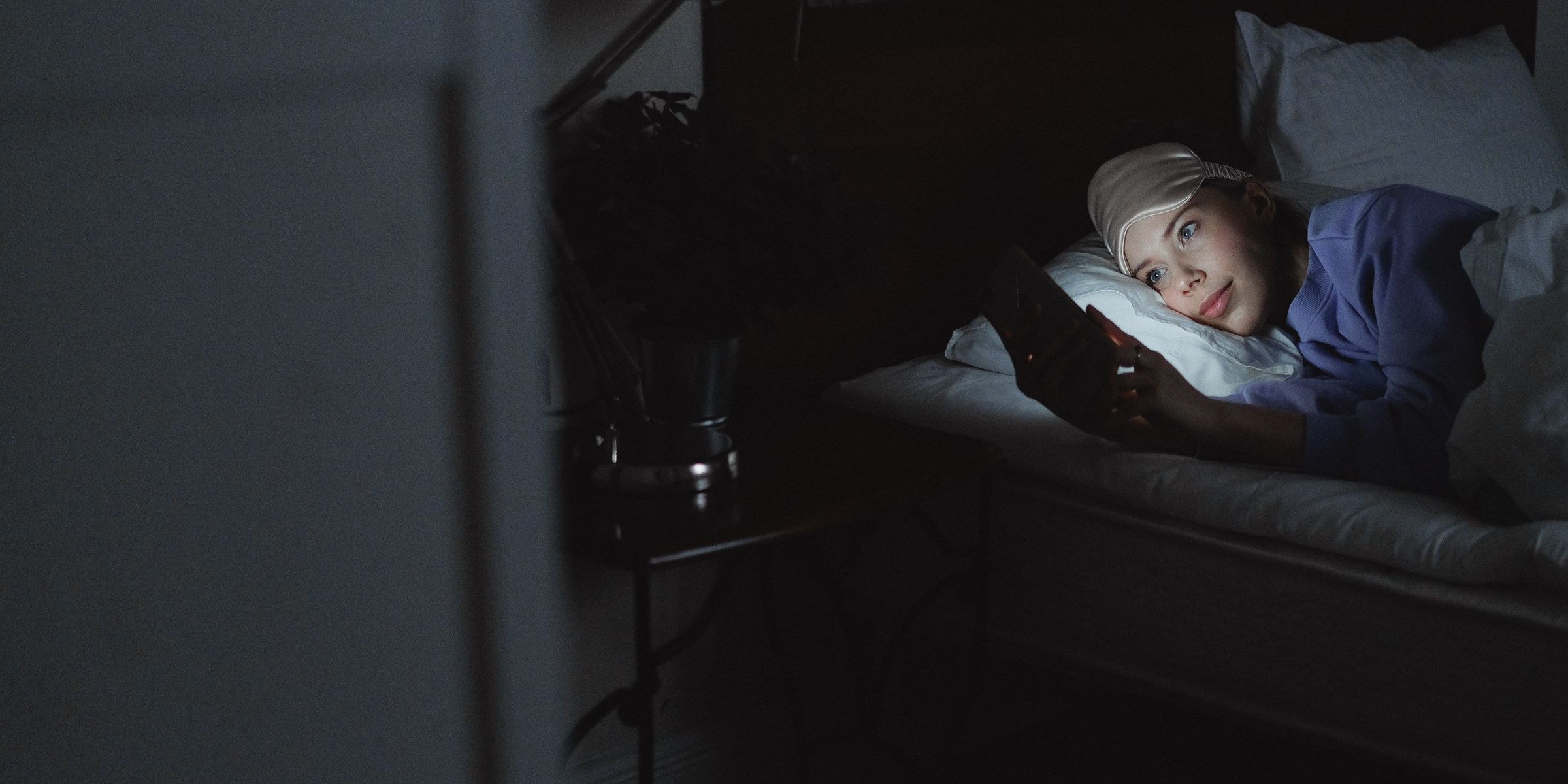 A young woman lying in bed at night, her face lit only by the glow of a phone screen